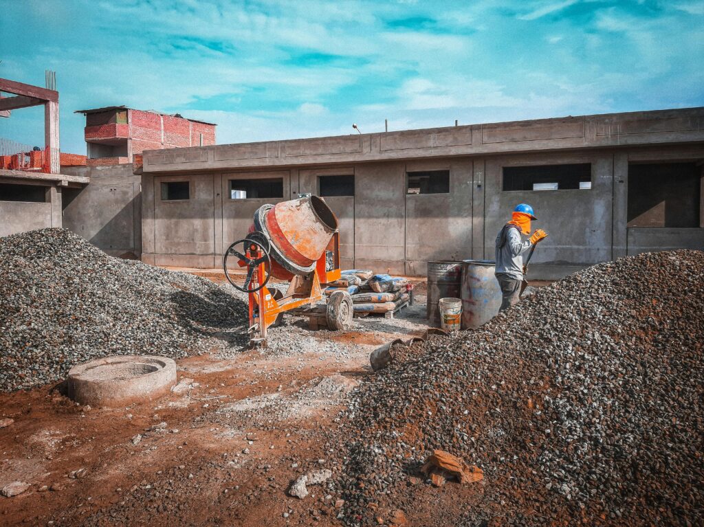 pexels photo 6034140 6034140 A construction site with a worker wearing safety gear next to a cement mixer and gravel piles.